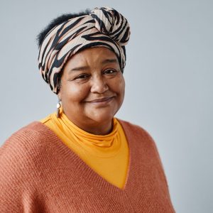 Mature woman wearing a patterned head scarf with gold hoop earrings and an orange jumper, smiling at the camera. She is standing in front of a plain grey background.