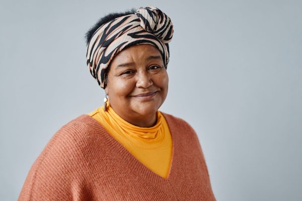 Mature woman wearing a patterned head scarf with gold hoop earrings and an orange jumper, smiling at the camera. She is standing in front of a plain grey background.