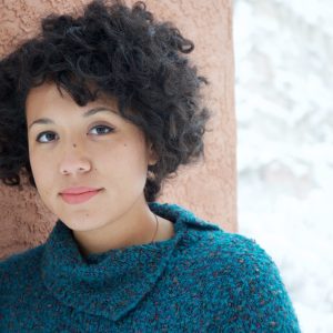 a young girl with big curly hair is standing against a wall, she is wearing a green jumper and has a neutral expression