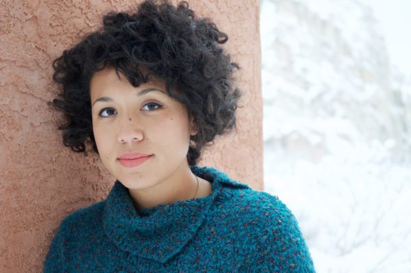 a young girl with big curly hair is standing against a wall, she is wearing a green jumper and has a neutral expression