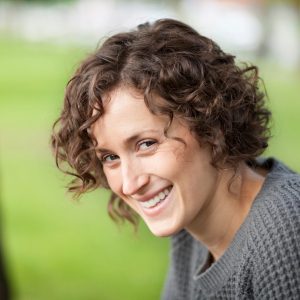 a young white women with long curly hair is looking to her left and smiling directly at the camera. The blurred background suggests she is in a green area, she is also wearing a grey jumper