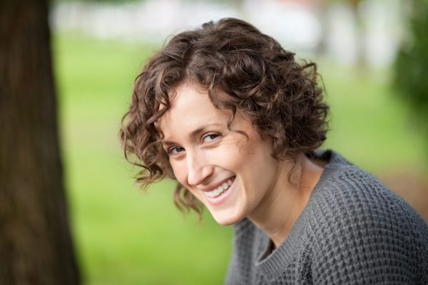 a young white women with long curly hair is looking to her left and smiling directly at the camera. The blurred background suggests she is in a green area, she is also wearing a grey jumper