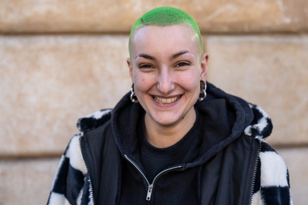 Young woman with bright green buzzcut hair. She has silver ear piercings and is wearing a black hoodie under a larger black and white coat. She is smiling widely, looking directly into the camera and is in front of a pale wall.