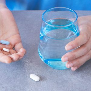 The photo shows a glass of water and medication, a pair of hands is holding the water and two tablets