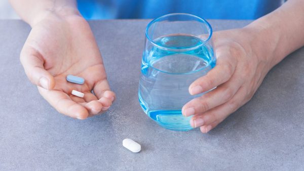 The photo shows a glass of water and medication, a pair of hands is holding the water and two tablets