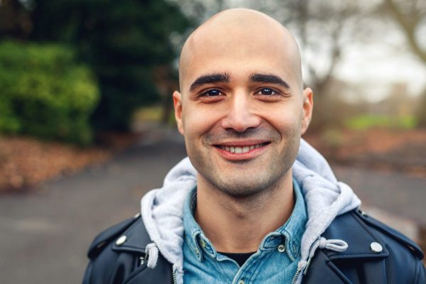 Young man who is bald, smiling directly at the camera. He is standing in a park with tree's behind him.