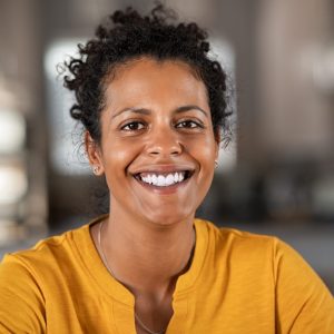 a women with short curly hair is smiling broadly at the camera, she is wearing a yellow jumper.