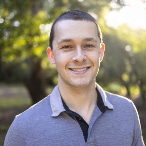 the photo shows a young man with short black hair and brown eyes standing outside and smiling straight at the camera