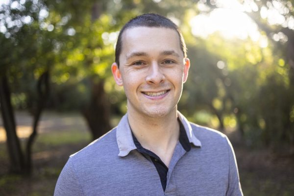 the photo shows a young man with short black hair and brown eyes standing outside and smiling straight at the camera