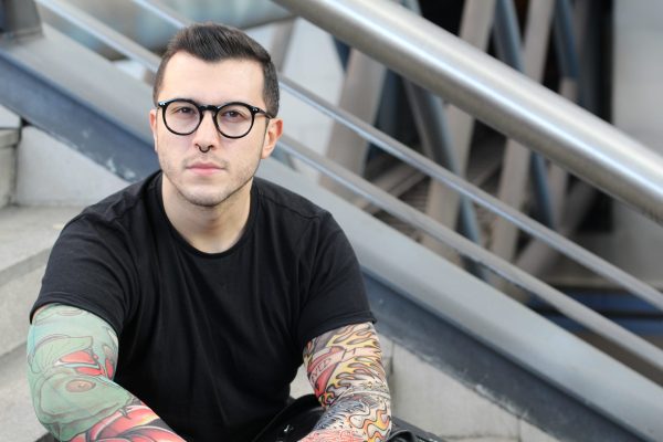 a young white man with short dark hair and glasses is sitting down on some stairs, he is wearing a black t shirt and looking at the camera with a neutral expression, he has tattoes on his arms.