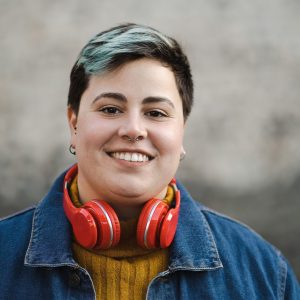 a young girl with short dark hair is smiling at the camera, her hair has green highlights and she has pierced ears and a nose piercing. She is wearing a jacket and has red headphones around her neck