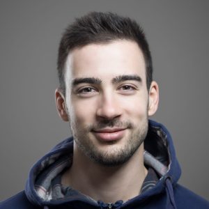 a young white man with short dark hair is smiling at the camera, he is also wearing a blue hoodie.