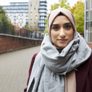 Young lady wearing a pale pink hijab and a grey scarf. She is looking into the camera with a slight smirk. She is standing in front of a brick wall and an apartment building in the background.