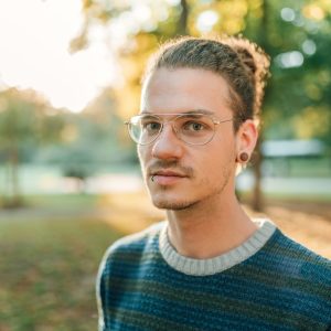 a young white man with short hair and slight facial hair and glasses is looking at the camera with a serious expression. He is wearing a green jumper and the blurred background appears to be a public park with trees.