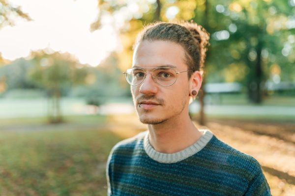 a young white man with short hair and slight facial hair and glasses is looking at the camera with a serious expression. He is wearing a green jumper and the blurred background appears to be a public park with trees.