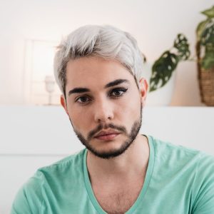 the photo shows a man with short white hair and brown eyes sitting inside and looking straight at the camera with a neutral expression