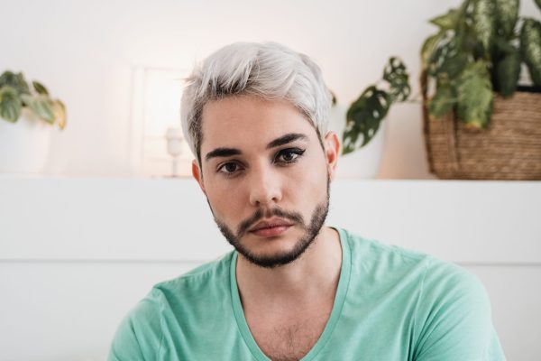 the photo shows a man with short white hair and brown eyes sitting inside and looking straight at the camera with a neutral expression