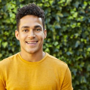 the photo shows a man with short curly black hair and brown eyes standing outside in front of bushes and smiling straight at the camera