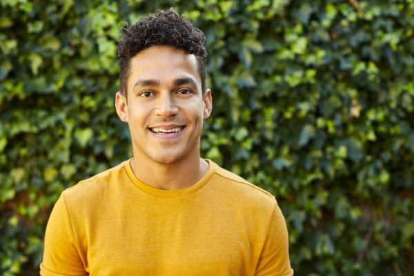 the photo shows a man with short curly black hair and brown eyes standing outside in front of bushes and smiling straight at the camera