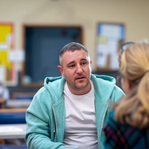 a man is sitting opposite a woman and talking, he has a serious expression and is wearing a light green hoodie