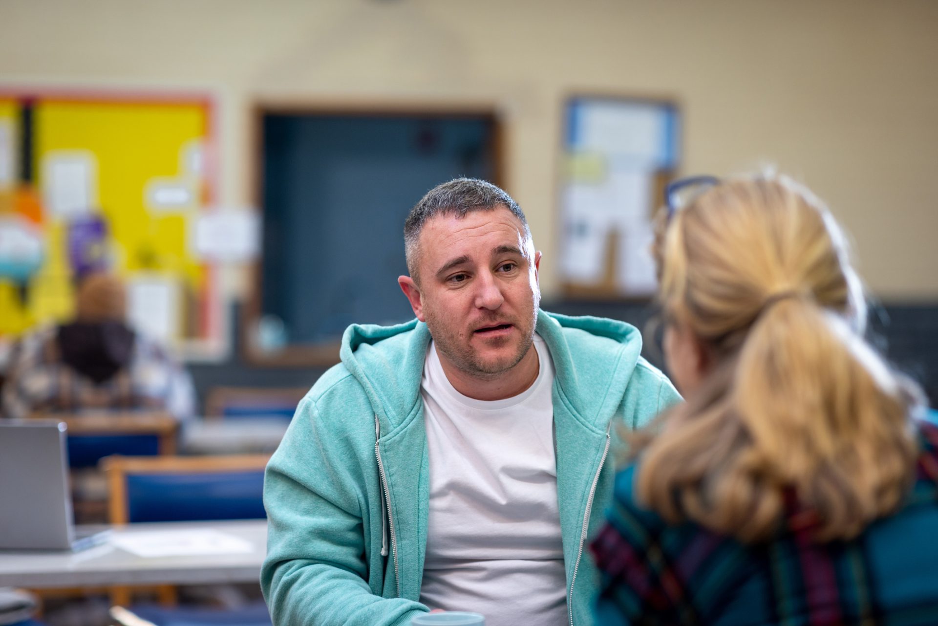 a man is sitting opposite a woman and talking, he has a serious expression and is wearing a light green hoodie