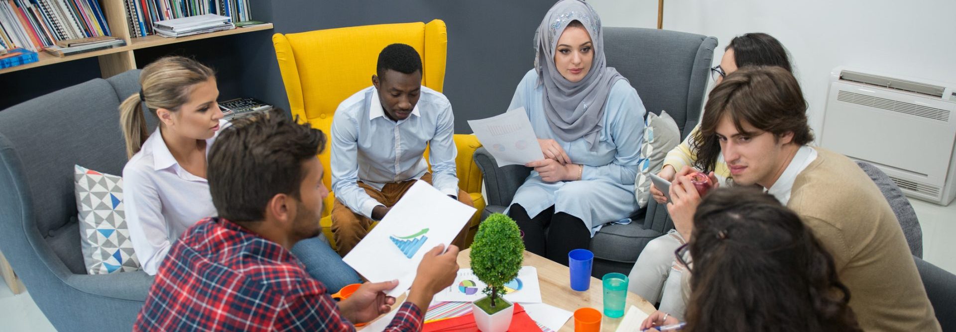 a group of people are sitting inside in a circle of armchairs and are engaged in a discussion, two of them are holding pieces of paper