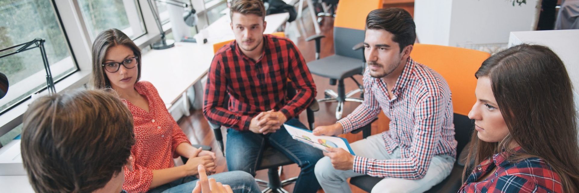 a group of people are sitting inside in a circle of chairs and are engaged in a discussion, one of them is holding a piece of paper that contains charts and graphs