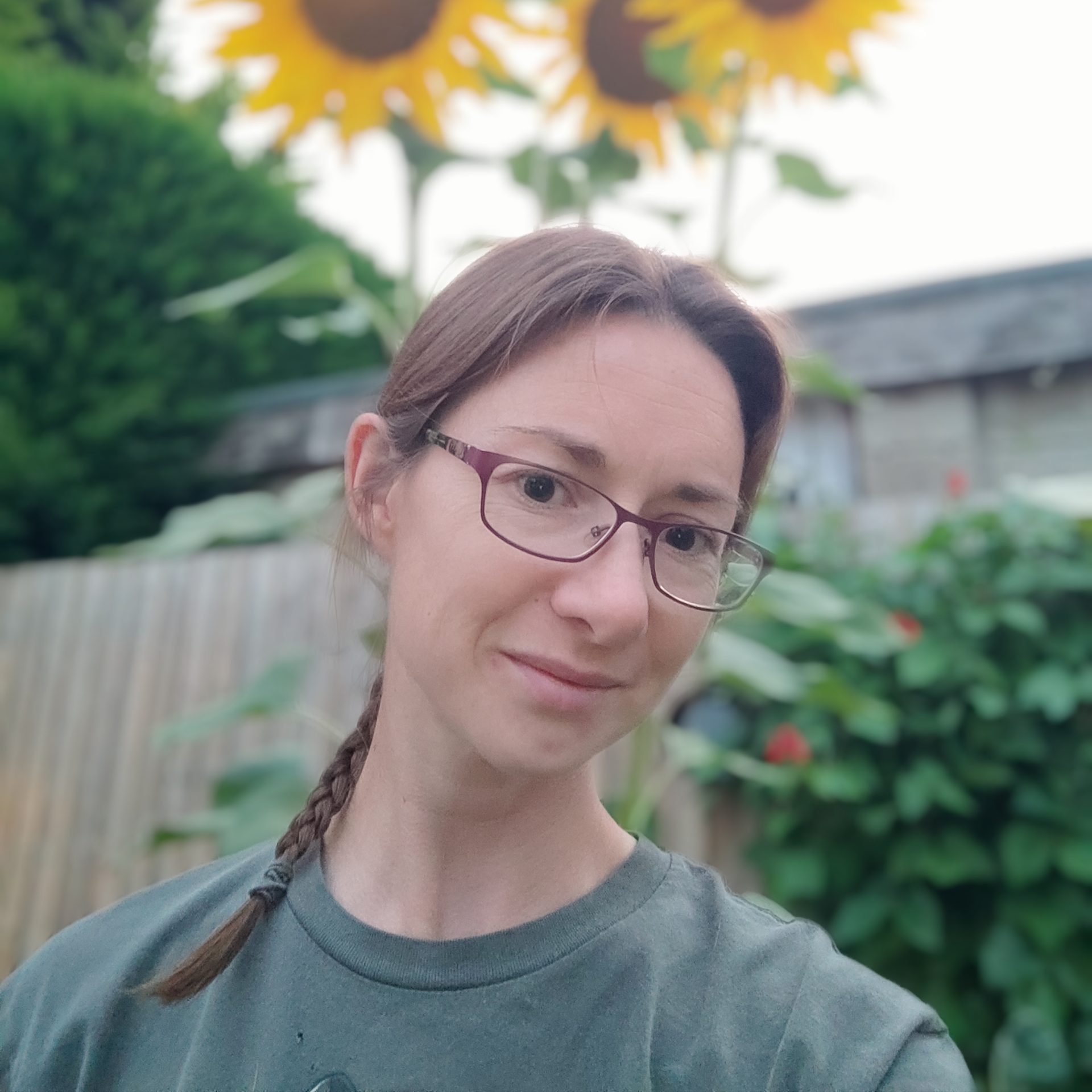 The photo shows our staff member Jenny standing outside, who has dark eyes and dark brown hair in a plat, she is wearing glasses and looking at the camera with a slight smile