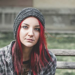 the photo shows a young girl sitting outside on a park bench, she has green eyes and long straight red hair, she is looking at the camera with a neutral expression