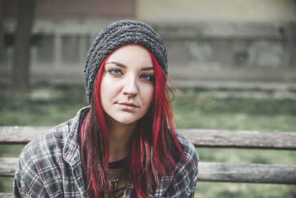 the photo shows a young girl sitting outside on a park bench, she has green eyes and long straight red hair, she is looking at the camera with a neutral expression