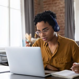 the photo shows a young woman who is wearing headphones and looking at her laptop screen while holding her pen above her note book