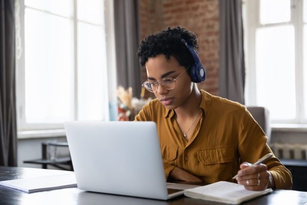 the photo shows a young woman who is wearing headphones and looking at her laptop screen while holding her pen above her note book