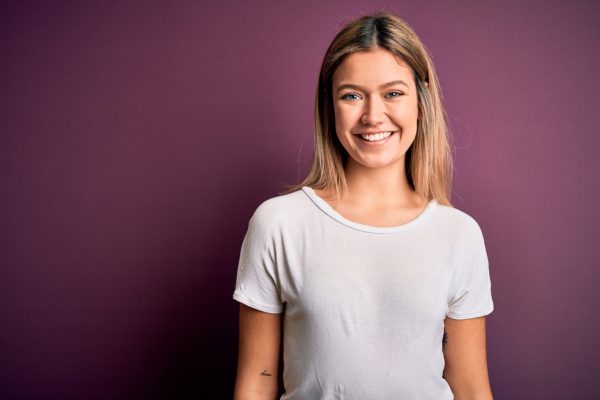 the photo shows a young woman standing in a room with a purple background, smiling straight at the camera