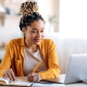 the photo shows a young woman who is looking at her laptop and holding a pen on top of her note book