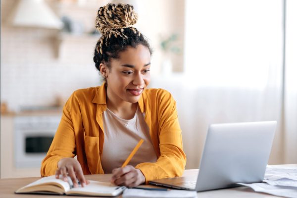 the photo shows a young woman who is looking at her laptop and holding a pen on top of her note book